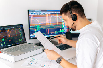 Successful male trader with headset sit at desk at home office monitoring stock market looking at...