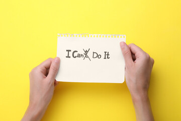Motivation concept. Woman holding paper with changed phrase from I Can't Do It into I Can Do It by crossing over letter T on yellow background, top view