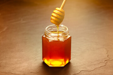 Natural honey dripping from dipper into glass jar on table under sunlight