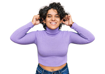 Young hispanic woman with curly hair wearing casual clothes smiling pulling ears with fingers,...
