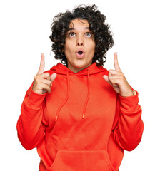 Young hispanic woman with curly hair wearing casual sweatshirt amazed and surprised looking up and pointing with fingers and raised arms.