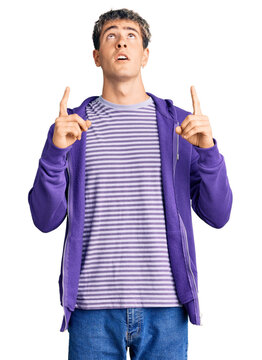 Young handsome man wearing casual purple sweatshirt amazed and surprised looking up and pointing with fingers and raised arms.