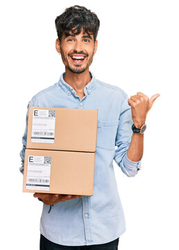 Young hispanic man holding delivery package pointing thumb up to the side smiling happy with open mouth
