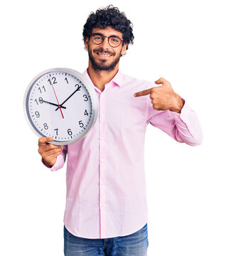Handsome young man with curly hair and bear holding big clock pointing finger to one self smiling happy and proud