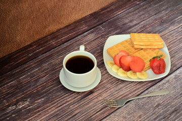 A plate with a bunch of fresh Viennese waffles with banana and strawberry slices on a wooden table, next to a cup of hot coffee.