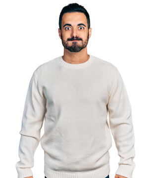 Young hispanic man with beard wearing casual white sweater puffing cheeks with funny face. mouth inflated with air, crazy expression.