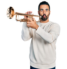 Young hispanic man with beard playing trumpet making fish face with mouth and squinting eyes, crazy and comical.