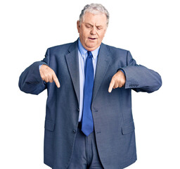 Senior grey-haired man wearing business jacket pointing down with fingers showing advertisement, surprised face and open mouth