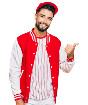 Young man with beard wearing baseball uniform smiling with happy face looking and pointing to the side with thumb up.