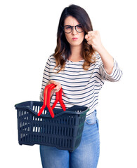 Beautiful young brunette woman holding supermarket shopping basket annoyed and frustrated shouting with anger, yelling crazy with anger and hand raised