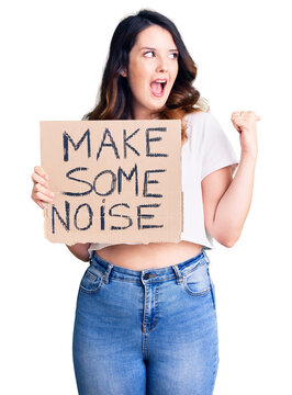 Beautiful young brunette woman holding make some noise banner pointing thumb up to the side smiling happy with open mouth