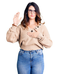 Beautiful young brunette woman wearing casual clothes and glasses swearing with hand on chest and...