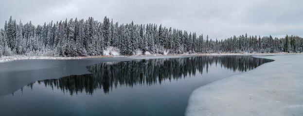 A panoramic view of a cold looking winter scene at a partially frozen lake that is surrounded by an evergreen forest. The trees are covered with heavy snow.  Tree reflections can be seen in the calm w