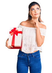 Young beautiful woman holding gift serious face thinking about question with hand on chin, thoughtful about confusing idea