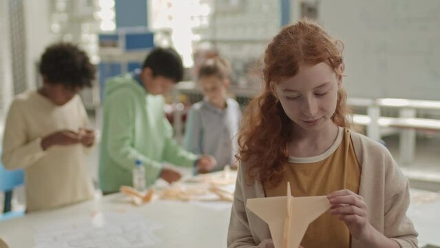 Waist up slowmo portrait of pretty ginger haired Caucasian schoolgirl holding DIY wooden plane in hands posing for camera in crafts classroom while her classmates woodworking in blurred background