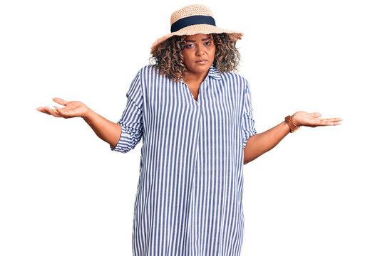 Young african american plus size woman wearing summer hat clueless and confused expression with arms and hands raised. doubt concept.
