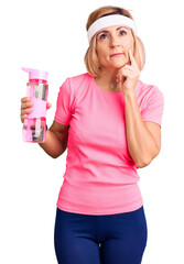 Young blonde woman wearing sportswear holding water bottle serious face thinking about question with hand on chin, thoughtful about confusing idea