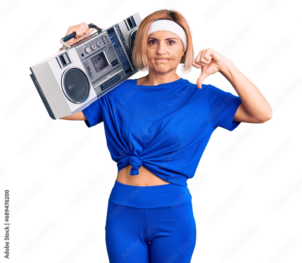 Sticker Young blonde woman wearing sportswear holding boombox, listening to music with angry face, negative sign showing dislike with thumbs down, rejection concept