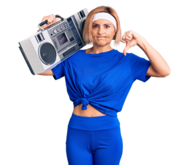Young blonde woman wearing sportswear holding boombox, listening to music with angry face, negative sign showing dislike with thumbs down, rejection concept