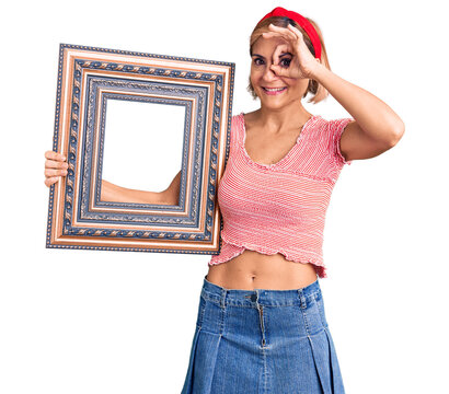 Young blonde woman holding empty frame smiling happy doing ok sign with hand on eye looking through fingers