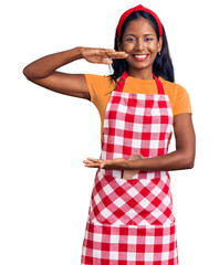 Young indian girl wearing professional baker apron gesturing with hands showing big and large size sign, measure symbol. smiling looking at the camera. measuring concept.
