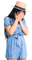 Brunette teenager girl wearing summer hat with sad expression covering face with hands while...