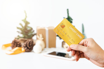 Woman's right hand holds golden credit card for online shopping on New Year's Day on background of Christmas tree, gift boxes, Christmas balls, pine cones and golden ribbon. There is a Clipping Path.