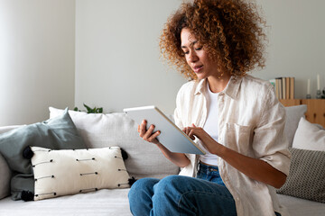 Young multiracial Hispanic woman using digital tablet sitting on the sofa at home living room. Copy...