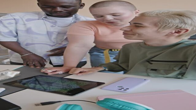 Vertical POV shot of positive multi-ethnic students typing programming code on digital tablet and speaking at camera while live streaming or video calling in college classroom