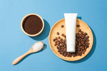 An unlabeled plastic tube and coffee beans on a wooden plate, coffee powder in a wooden bowl and a spoonful of sugar on a green background. Exfoliate with natural ingredients.