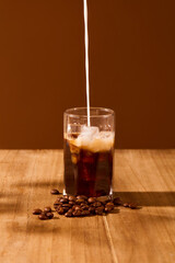 A stream of milk is pouring from above into a cup of black coffee on a wooden table with a brown background. Art space for food and beverage advertising.