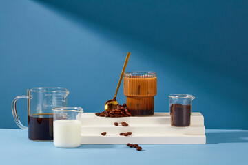 A glass of milky coffee is placed on a white platform with a metal spoon and coffee beans. Black...