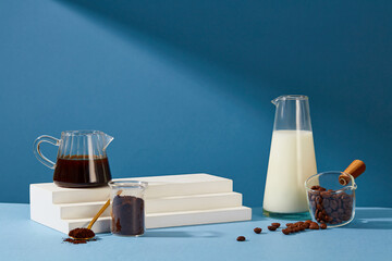 Black coffee, fresh milk, coffee powder, white podium and coffee beans are displayed on a blue background. Coffee also acts as an exfoliant to help reduce premature aging of the skin.