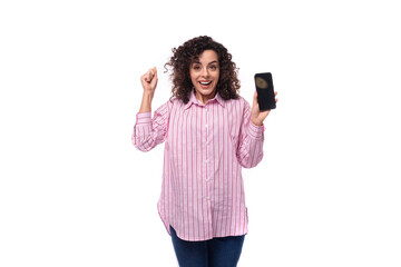young curly caucasian woman dressed in a striped pink shirt rejoices holding a phone with a mockup