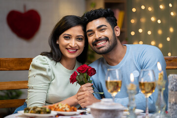 Happy young couple holding red rose by looking at camera during candlelight dinner at home - concept of valentines day, togetherness and dating