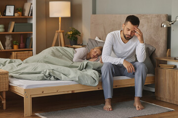 Sad man sitting on bed with female partner sleeping in background