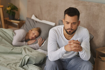 Sad man sitting on bed with female partner sleeping in background