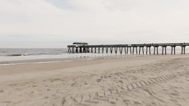 Drone of an empty beach in Tybee Island approaching pier and ocean with rough waves, low to the sand