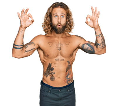 Handsome man with beard and long hair standing shirtless showing tattoos looking surprised and shocked doing ok approval symbol with fingers. crazy expression