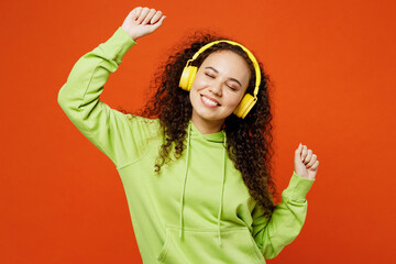 Young cheerful fun cool smiling woman of African American ethnicity she wear green hoody casual clothes listen to music in headphones dance isolated on plain red orange background. Lifestyle concept.