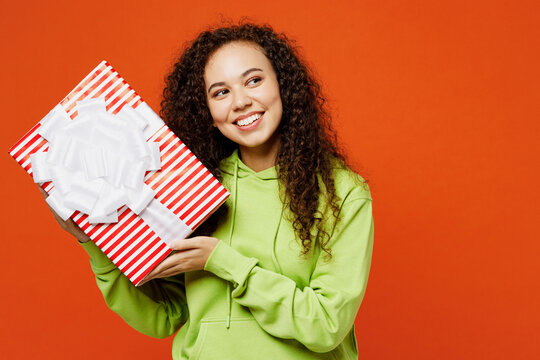 Young fun woman of African American ethnicity she wear green hoody casual clothes hold present box with gift ribbon bow look aside on area isolated on plain red orange background. Lifestyle concept.