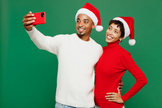 Merry young couple friends man woman wear red casual clothes Santa hat posing doing selfie shot on mobile cell phone isolated on plain green background. Happy New Year 2024 Christmas holiday concept.