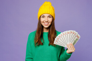 Young fun woman she wear green sweater yellow hat casual clothes hold in hand fan of cash money in dollar banknotes isolated on plain pastel light purple background studio portrait. Lifestyle concept.