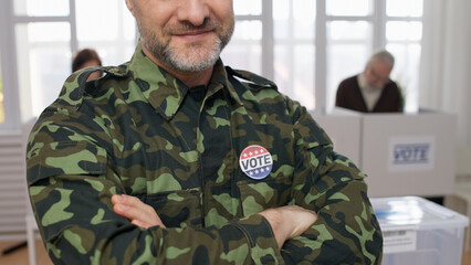 A smiling soldier in uniform with a voting pin standing at the polling place, folding his hands on...