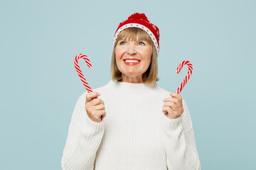 Merry minded elderly woman 50s years old wear sweater red hat posing hold hand candy cane lollipop look aside isolated on plain blue background. Happy New Year celebration Christmas holiday concept.