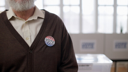 A close-up of a senior man with a vote button on his chest standing in the hall during the...