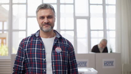 A middle-aged man showing off a vote pin on his chest at the polling station, citizen's responsibility