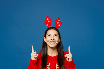 Merry smiling young Latin woman in red Christmas sweater decorative fun deer horns on head posing...