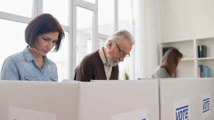 Concentrated men and women are reading information on election ballots before casting their votes