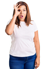 Fototapeta premium Middle age latin woman wearing casual white tshirt doing ok gesture shocked with surprised face, eye looking through fingers. unbelieving expression.